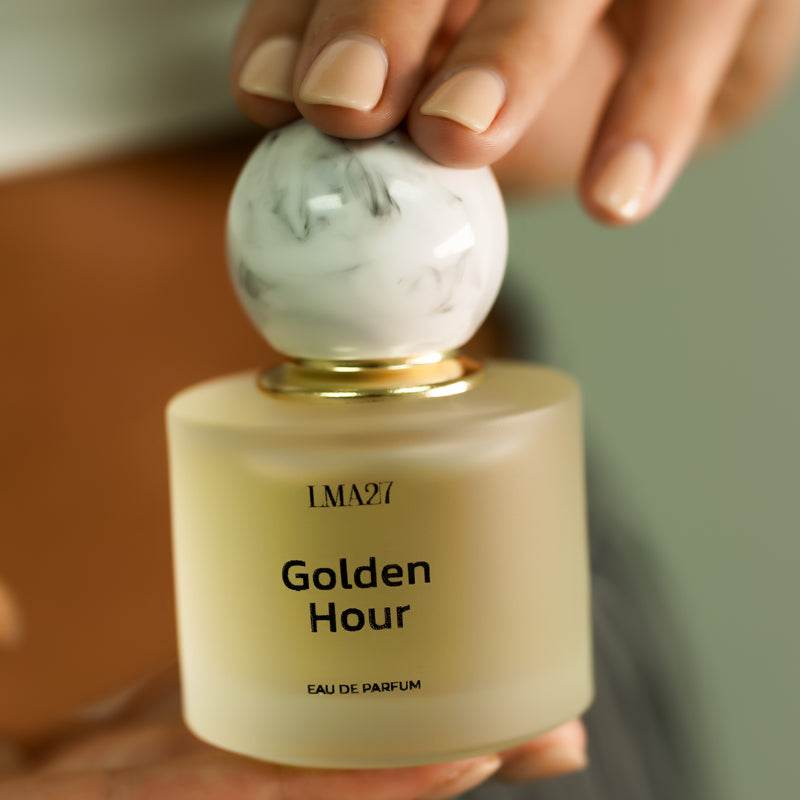 A hand holding a bottle of LMA27 Golden Hour Eau de Parfum. The perfume features a frosted glass bottle with a minimalist black font and a marble-textured round cap with a gold accent.
