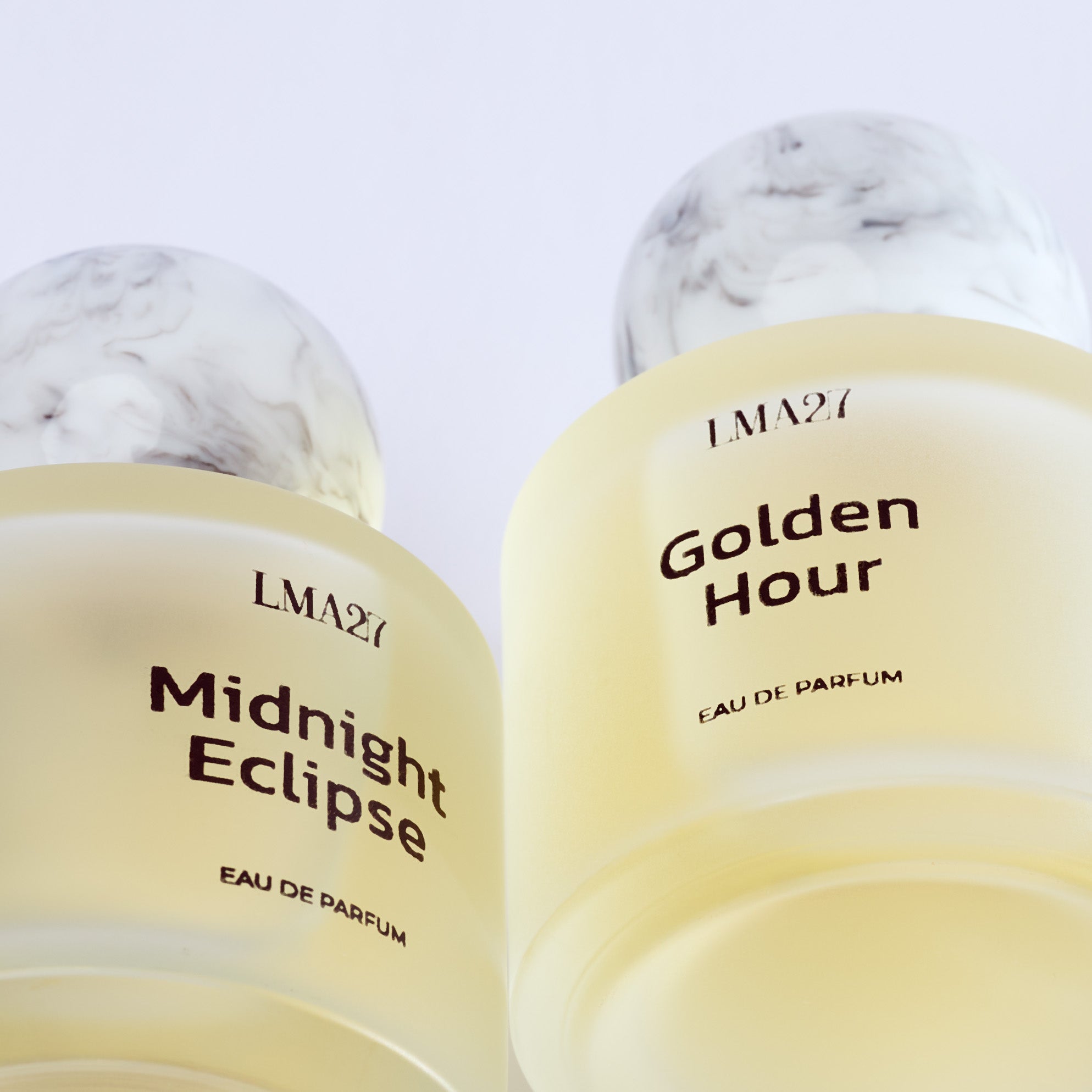 Two elegant luxury perfume bottles from brand LMA27, named "Midnight Eclipse" and "Golden Hour," featuring frosted glass with marble-textured spherical caps and minimalist black text labels, photographed on sky view.