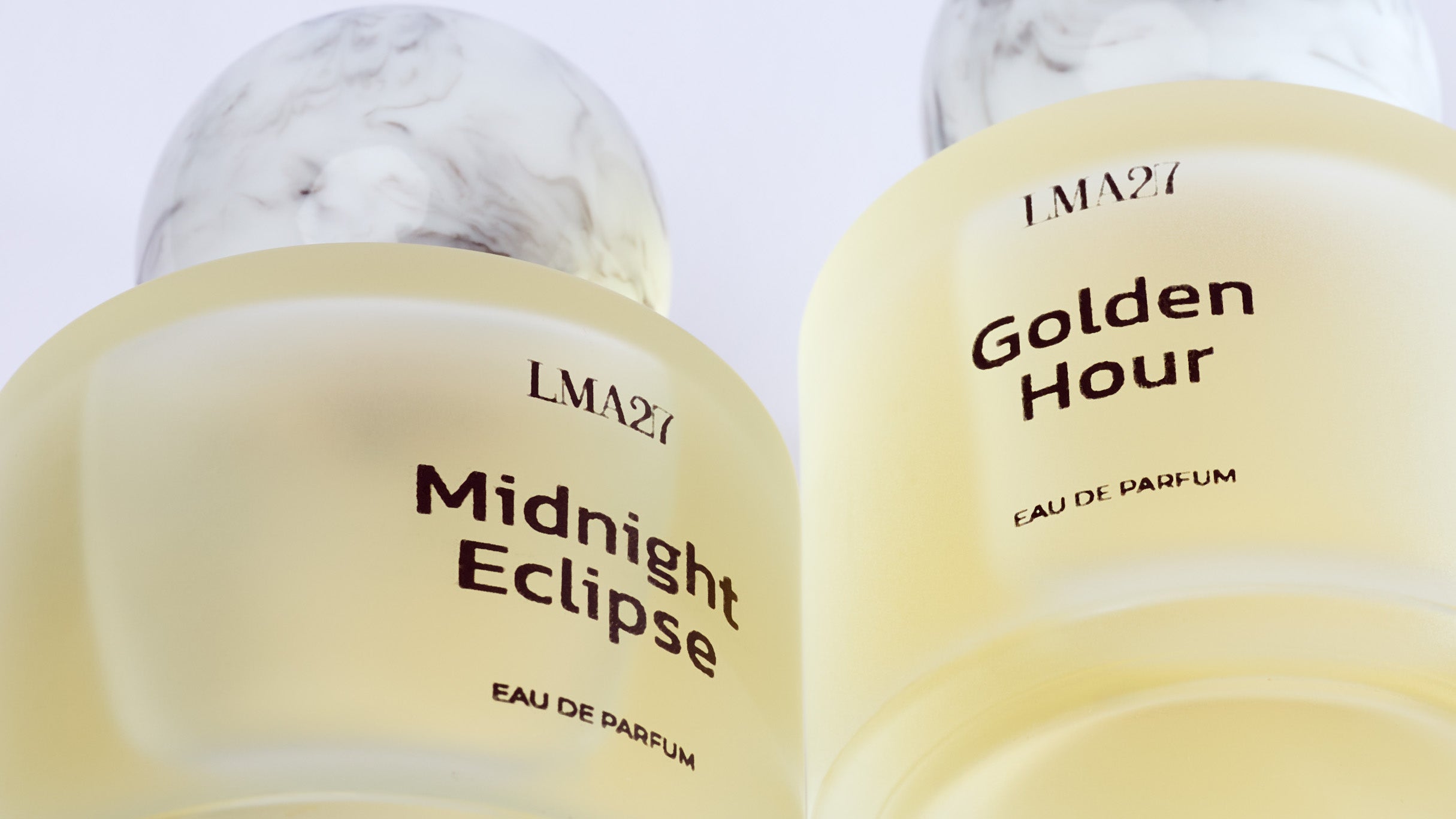 Two elegant luxury perfume bottles from brand LMA27, named "Midnight Eclipse" and "Golden Hour," featuring frosted glass with marble-textured spherical caps and minimalist black text labels, photographed on sky view.