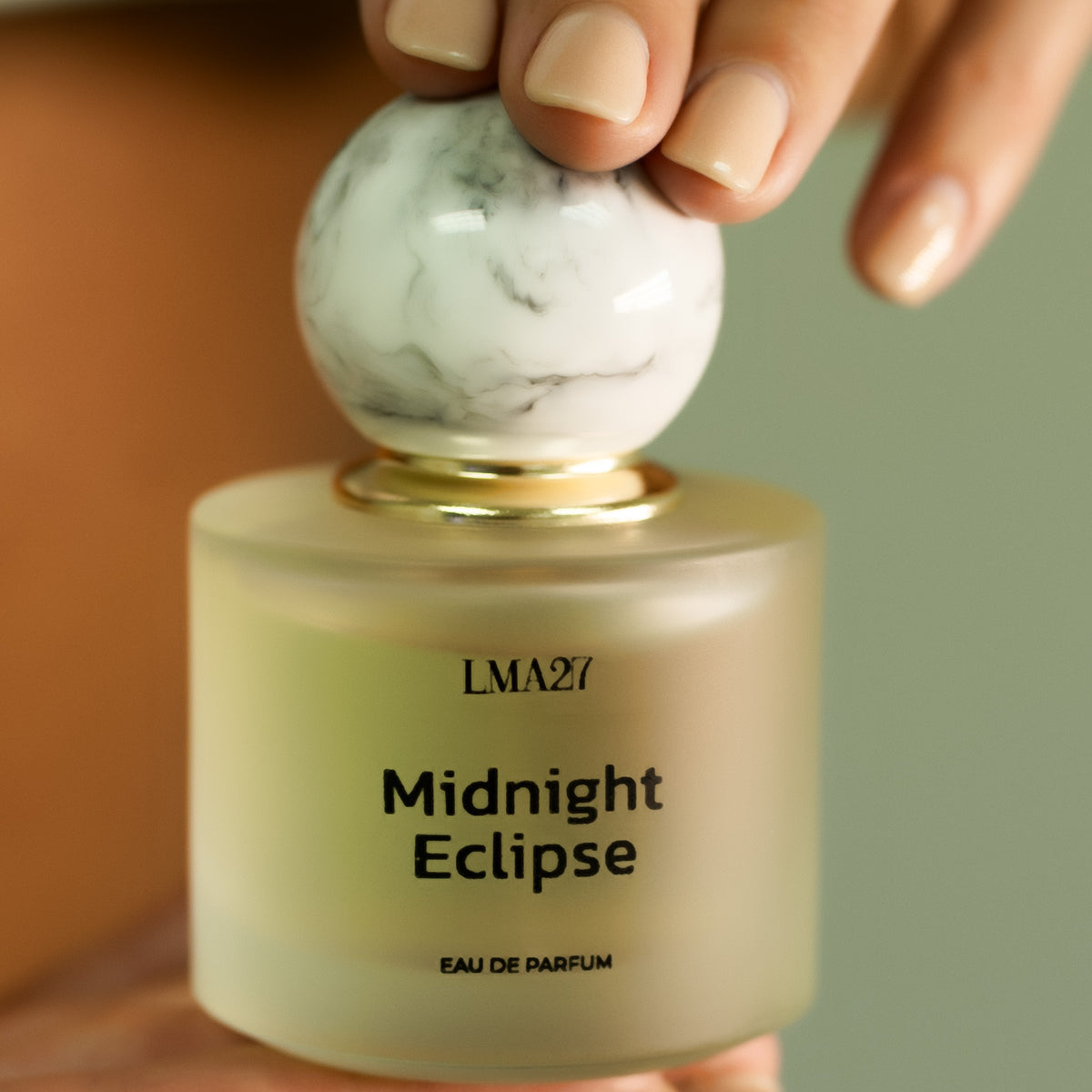 A hand holding a bottle of LMA27 MIDNIGHT ECLIPSE Eau de Parfum. The perfume features a frosted glass bottle with a minimalist black font and a marble-textured round cap with a gold accent.