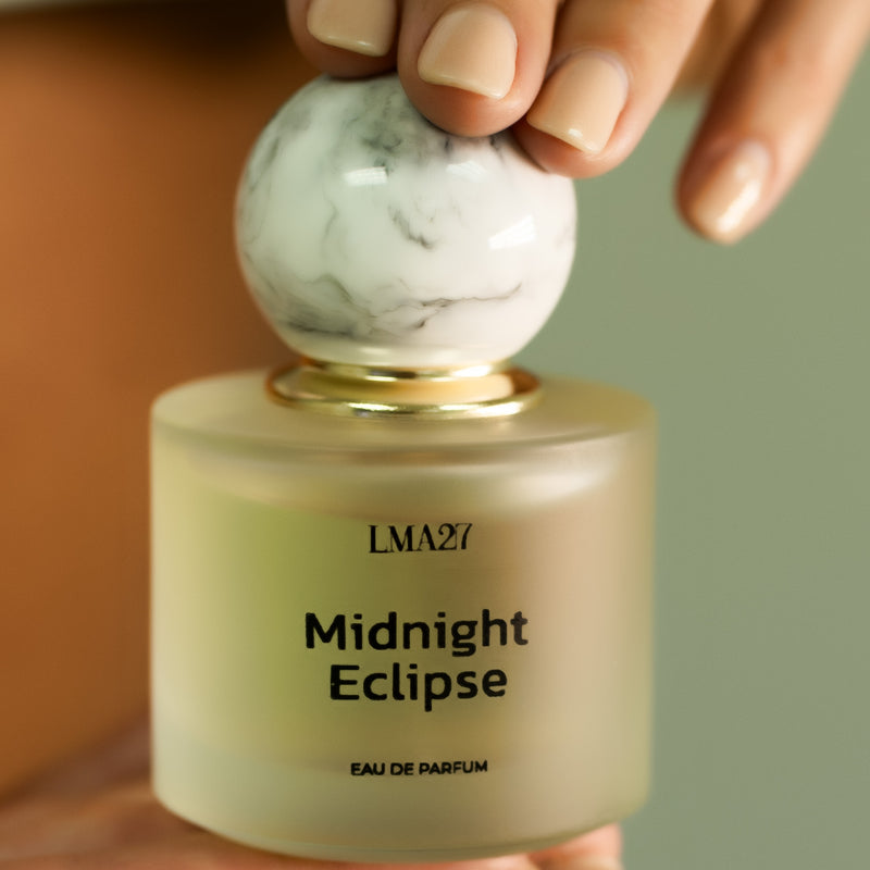 A hand holding a bottle of LMA27 MIDNIGHT ECLIPSE Eau de Parfum. The perfume features a frosted glass bottle with a minimalist black font and a marble-textured round cap with a gold accent.