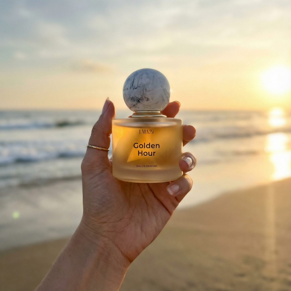 Hand holding a 'Golden Hour' perfume bottle with a sunset beach background